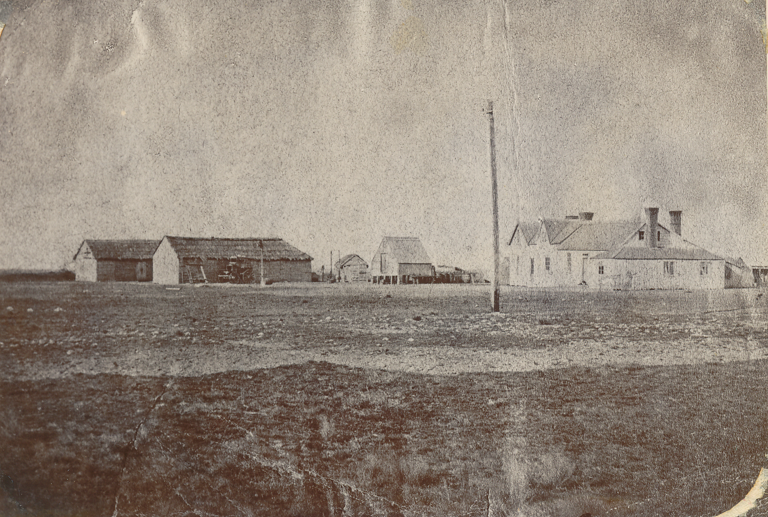 Early view of the Ashburton Arms accommodation house. Sepia tone 1850s - 1860s photo. Barren landscape, stables buildings at left, small hut in middle, flagpole piddle distance, inn at right of frame.